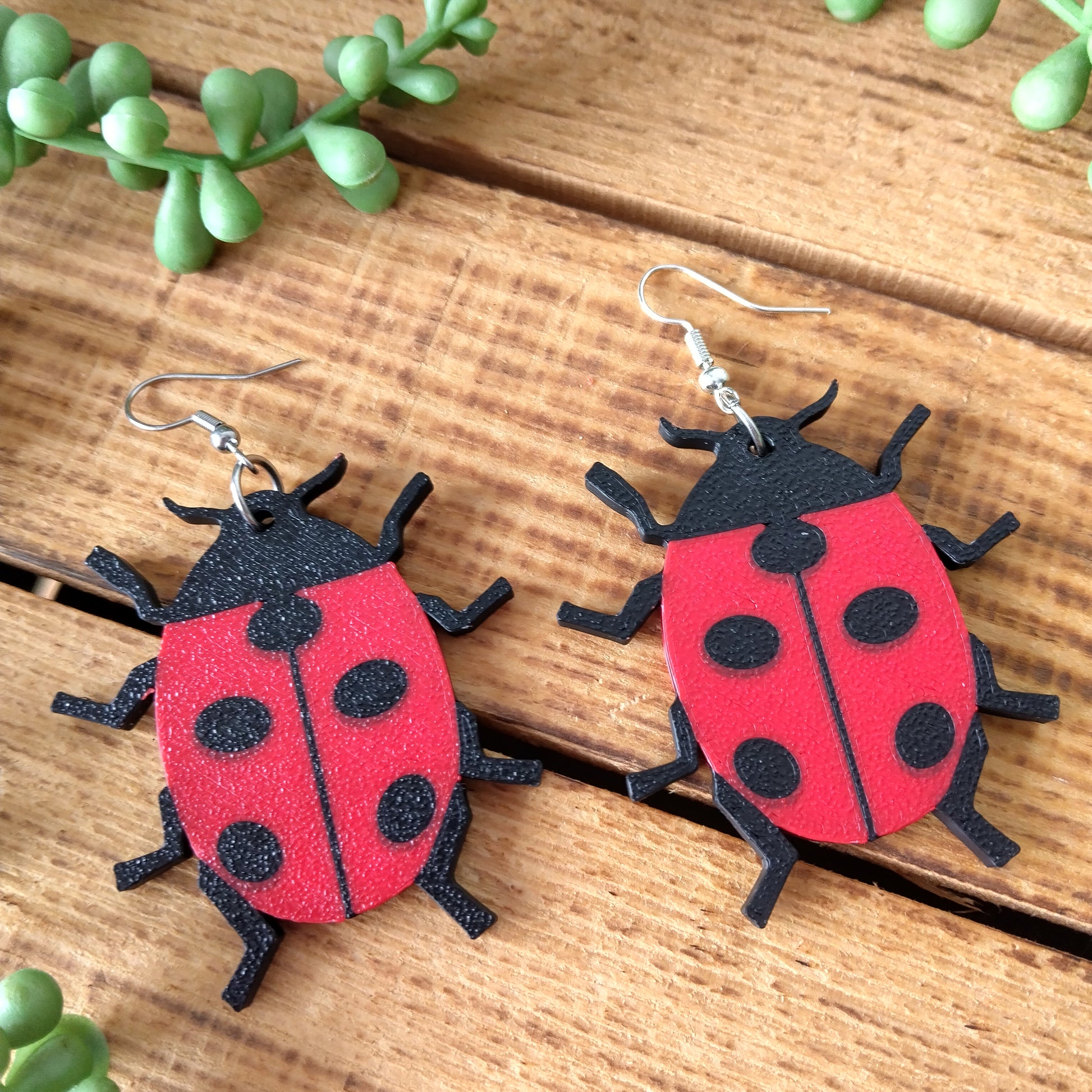 Red and black ladybug-shaped earrings on a wooden surface with green plants.