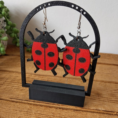 3d printed red and black ladybird-shaped earrings on a black stand with a wooden surface background. 
